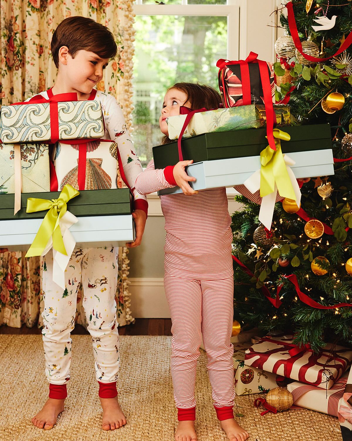 A cheerful brother and sister in matching pajamas carry stacks of wrapped presents in front of a decorated Christmas tree indoors.
