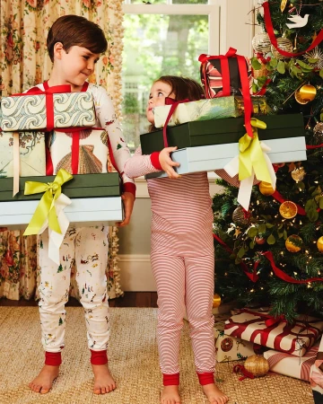 A cheerful brother and sister in matching pajamas carry stacks of wrapped presents in front of a decorated Christmas tree indoors.