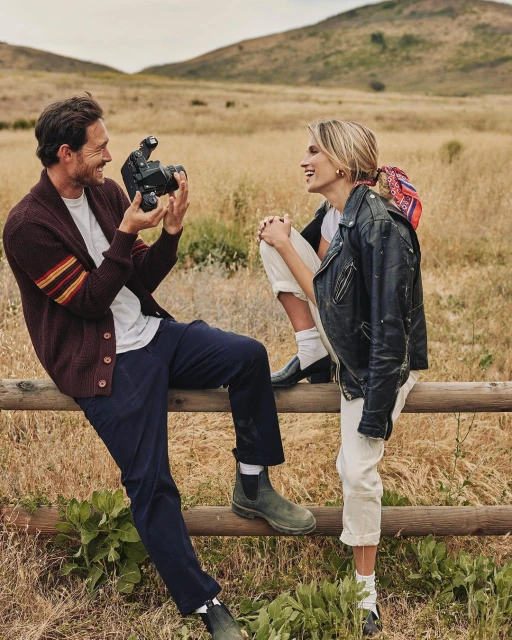 A man photographs a woman sitting on a wooden fence in a grassy field with rolling hills in the background, on a sunny day.