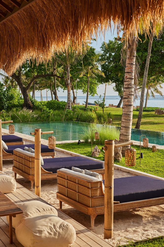 Outdoor luxury resort with lounge chairs near a pool and view of the ocean, shaded by a thatched roof and palm trees.