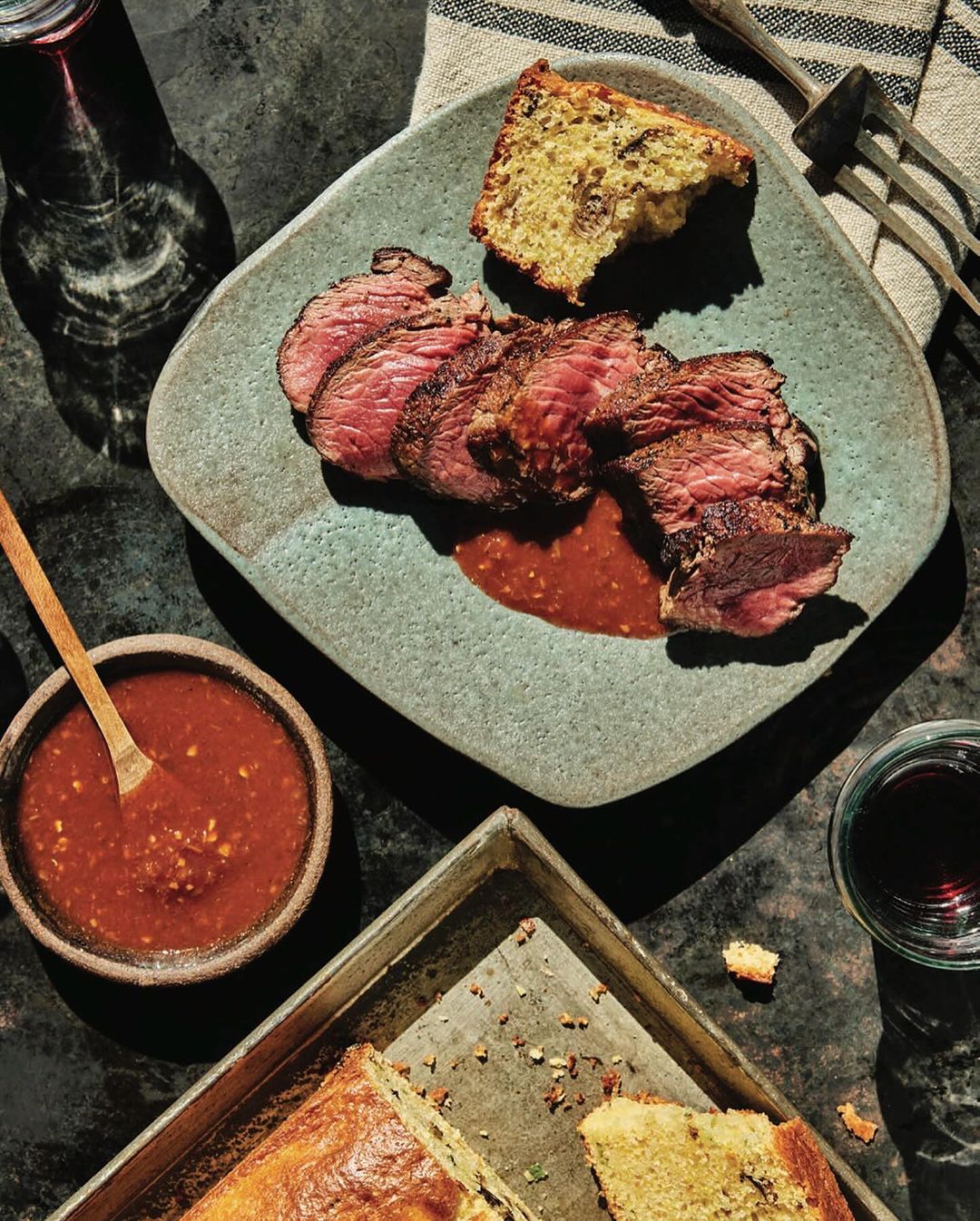 Overhead shot of sliced steak and cornbread served on a blue plate with a side of red sauce and a drink on a dark surface.