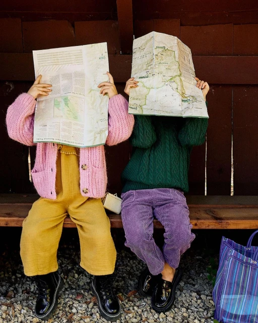 Two children sit, maps in front of their faces, wearing colorful clothes and black shoes, in an outdoor setting with a brown backdrop.
