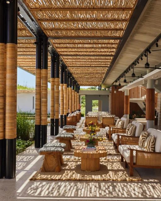 Indoor lounge area with rattan furniture, wood benches, and tropical flowers. Sunlight streams through the open-air ceiling.