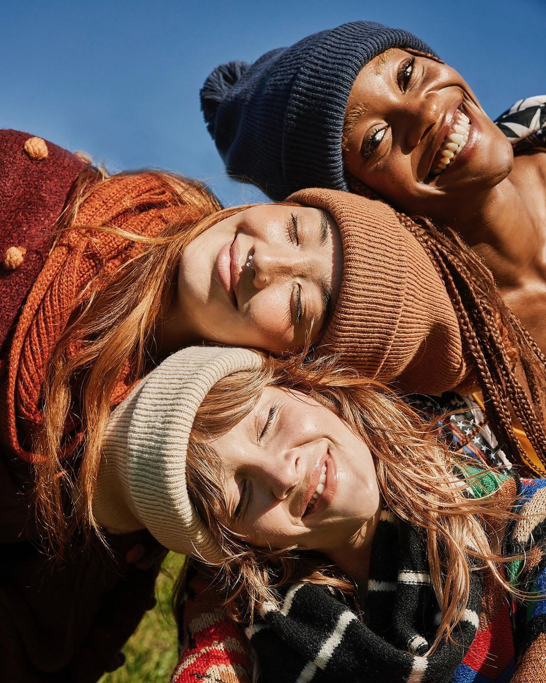 Eye-level close-up shot of three smiling women wearing knit caps in an outdoor setting, with the blue sky visible in the background.