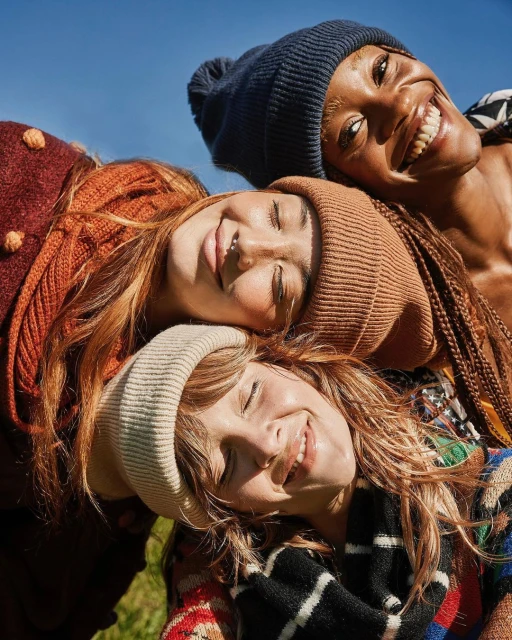 Eye-level close-up shot of three smiling women wearing knit caps in an outdoor setting, with the blue sky visible in the background.