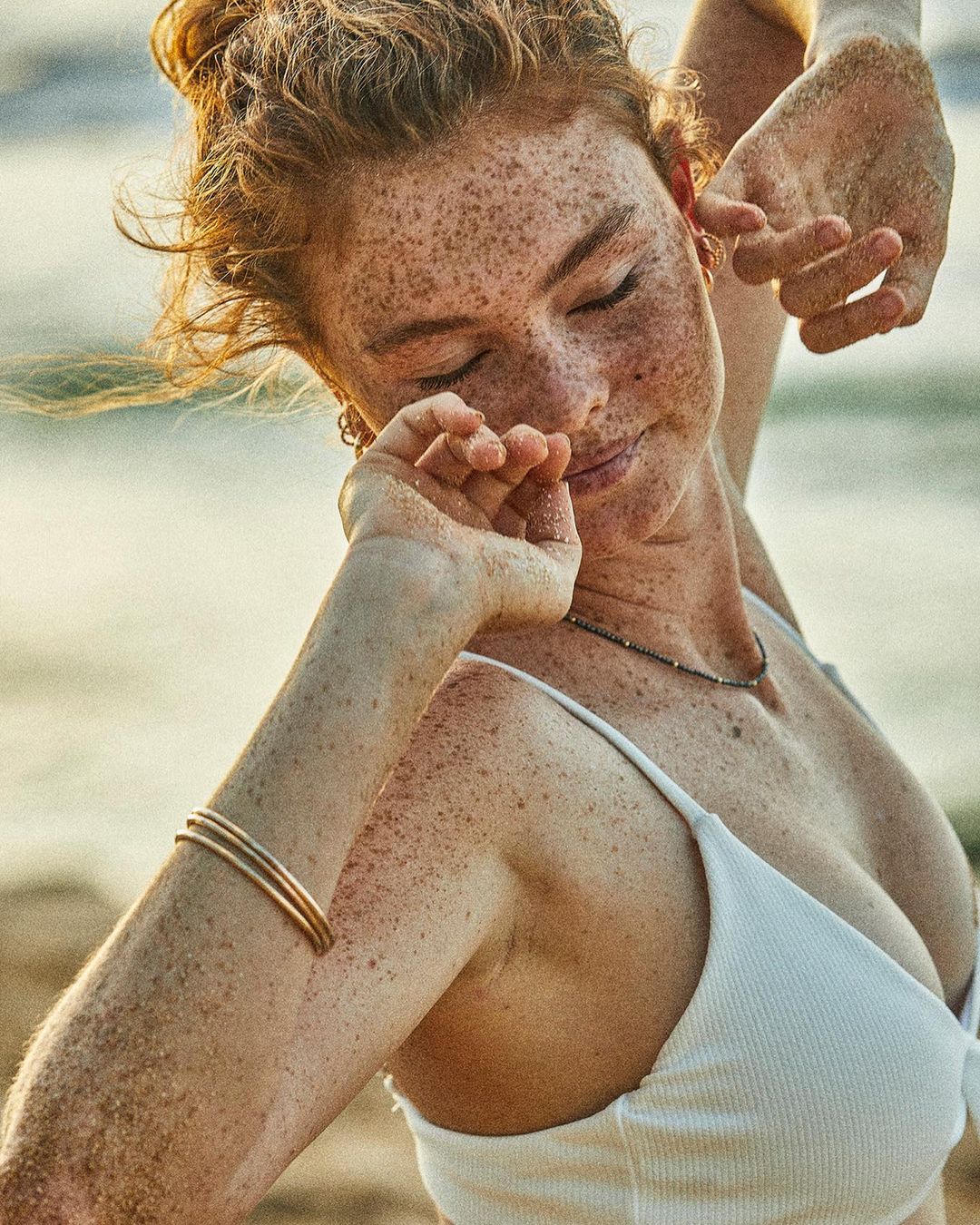 Close-up of a freckled woman in a white top at the beach with her eyes closed, arm raised, and sand on her hands.