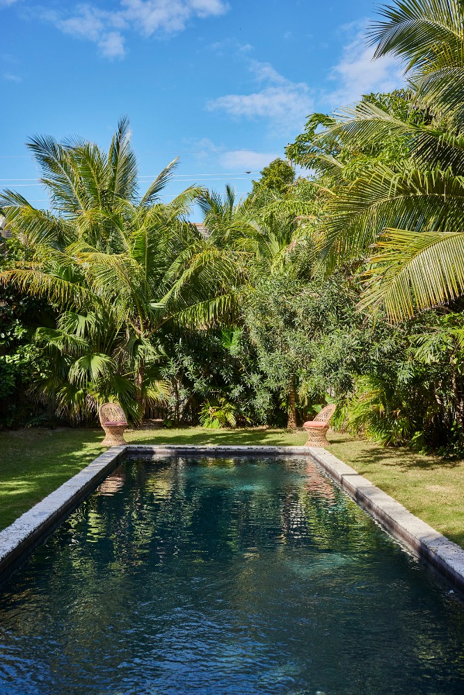 Outdoor swimming pool with dark water surrounded by a stone border and lush green tropical trees and blue sky.