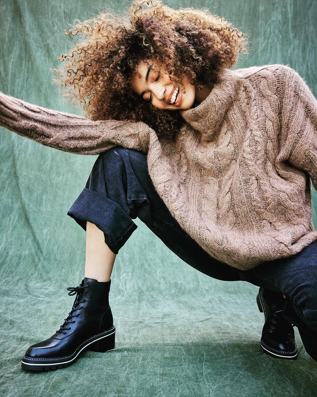 Smiling woman with curly hair in a light brown sweater and black boots poses in front of a mottled light-green background in a fashion shot.