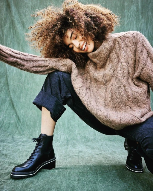 Smiling woman with curly hair in a light brown sweater and black boots poses in front of a mottled light-green background in a fashion shot.
