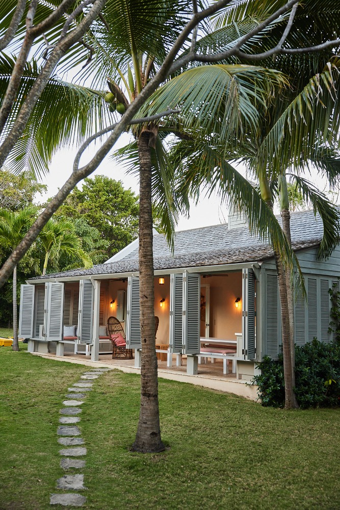 Exterior view of a light grey house with open shutters, surrounded by lush green grass, tropical palm trees, and a stone walkway.
