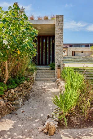 Sandy pathway leading to a modern stone and concrete building entrance framed by lush green foliage under a blue sky.
