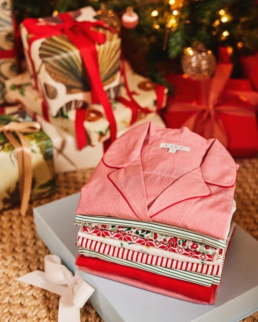 Stack of folded pajamas on a gift box under a Christmas tree with festive wrapped presents, creating a cozy holiday atmosphere.