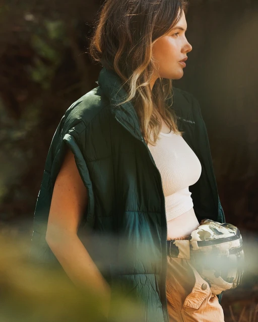 A young woman with medium brown hair is outdoors wearing a green vest and beige pants, looking toward the right in soft light.