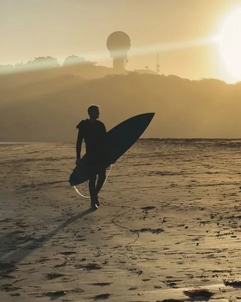 Silhouette of a surfer walking on a sandy beach with a surfboard at sunset, with a radar tower visible on a distant hill.