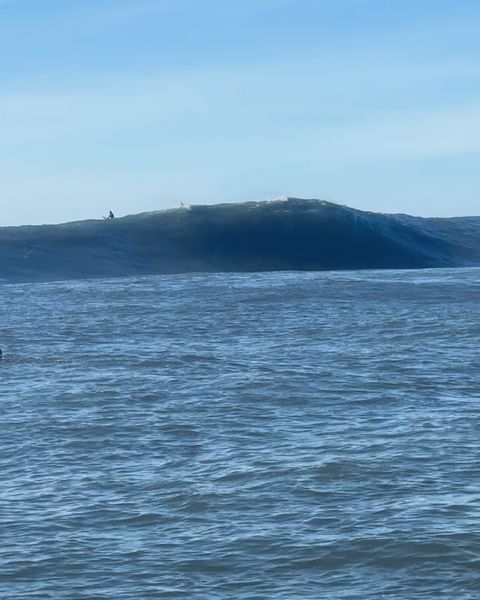 Ocean wave cresting with white water; dark blue sea under light blue sky; surfer visible far off on the wave crest.