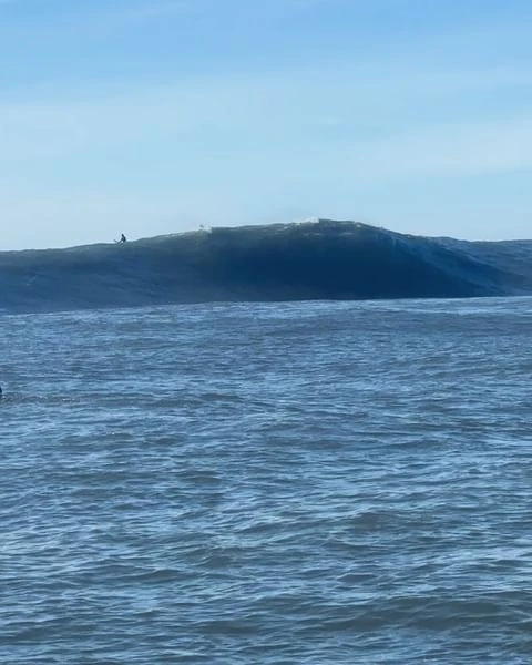 Ocean wave cresting with white water; dark blue sea under light blue sky; surfer visible far off on the wave crest.