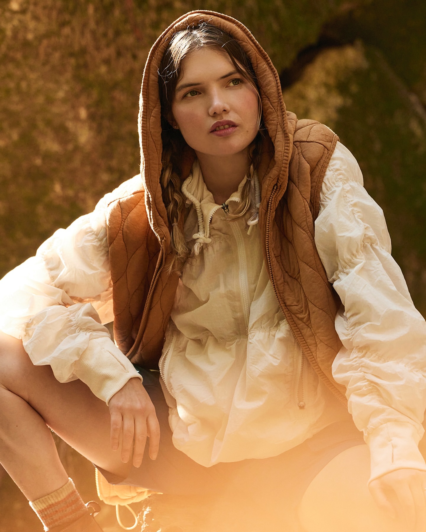 Young woman in a quilted vest with a hood crouching in a forest with warm, diffused sunlight shining through the trees.