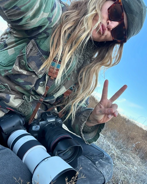 Smiling woman in camouflage jacket poses outdoors giving the peace sign with camera equipment and lens while wearing sunglasses and a beanie.