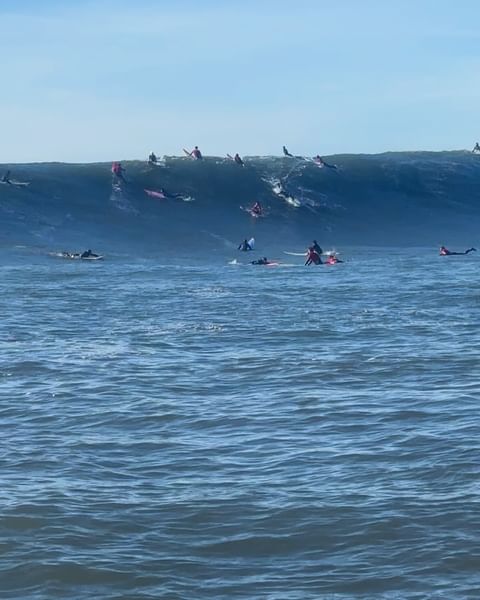 A group of surfers on surfboards waiting to ride a large ocean wave on a clear day under a blue sky, showcasing the sport of surfing.