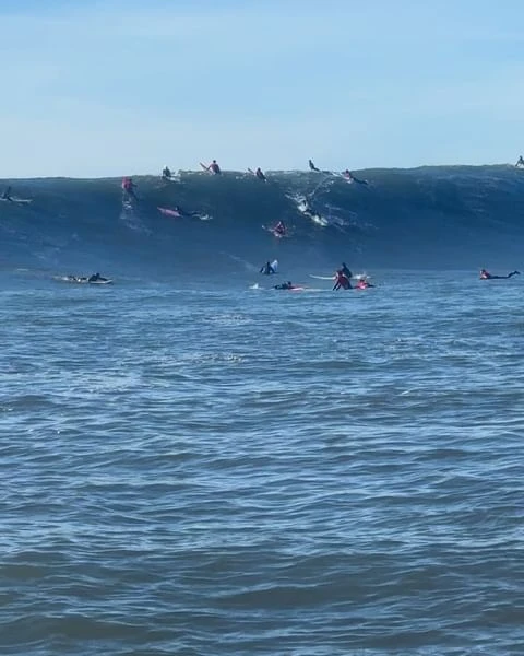 A group of surfers on surfboards waiting to ride a large ocean wave on a clear day under a blue sky, showcasing the sport of surfing.