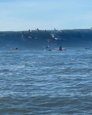 A group of surfers on surfboards waiting to ride a large ocean wave on a clear day under a blue sky, showcasing the sport of surfing.