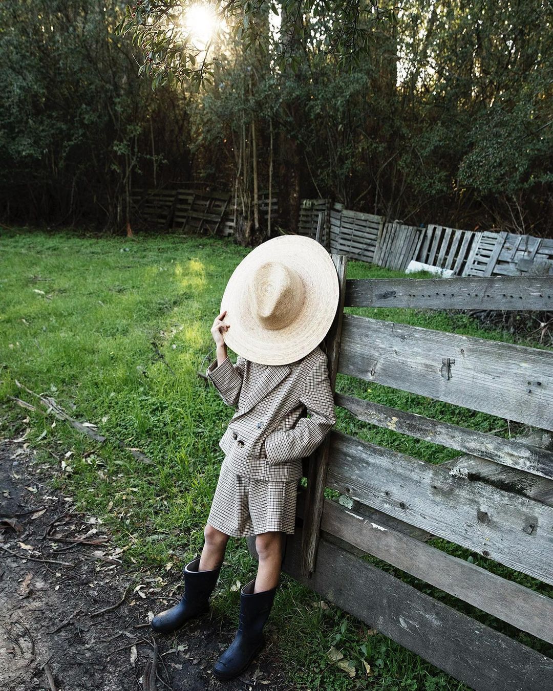 Child in oversized straw hat and houndstooth suit leans against weathered wooden fence in a sun-drenched, lush green outdoor setting.