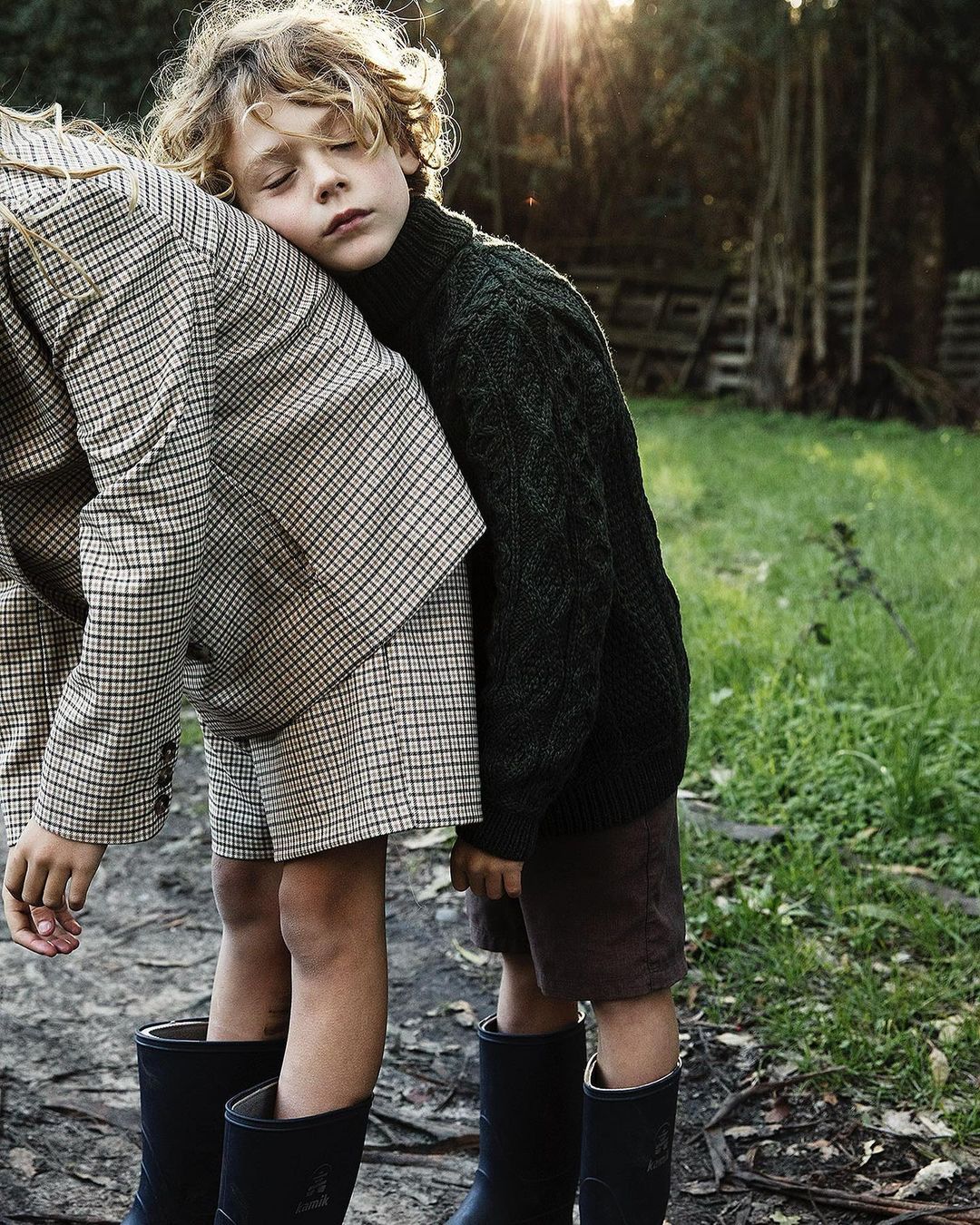 Two young children stand in a grassy field, one with blonde curly hair resting their head on the other's shoulder in the sunshine.