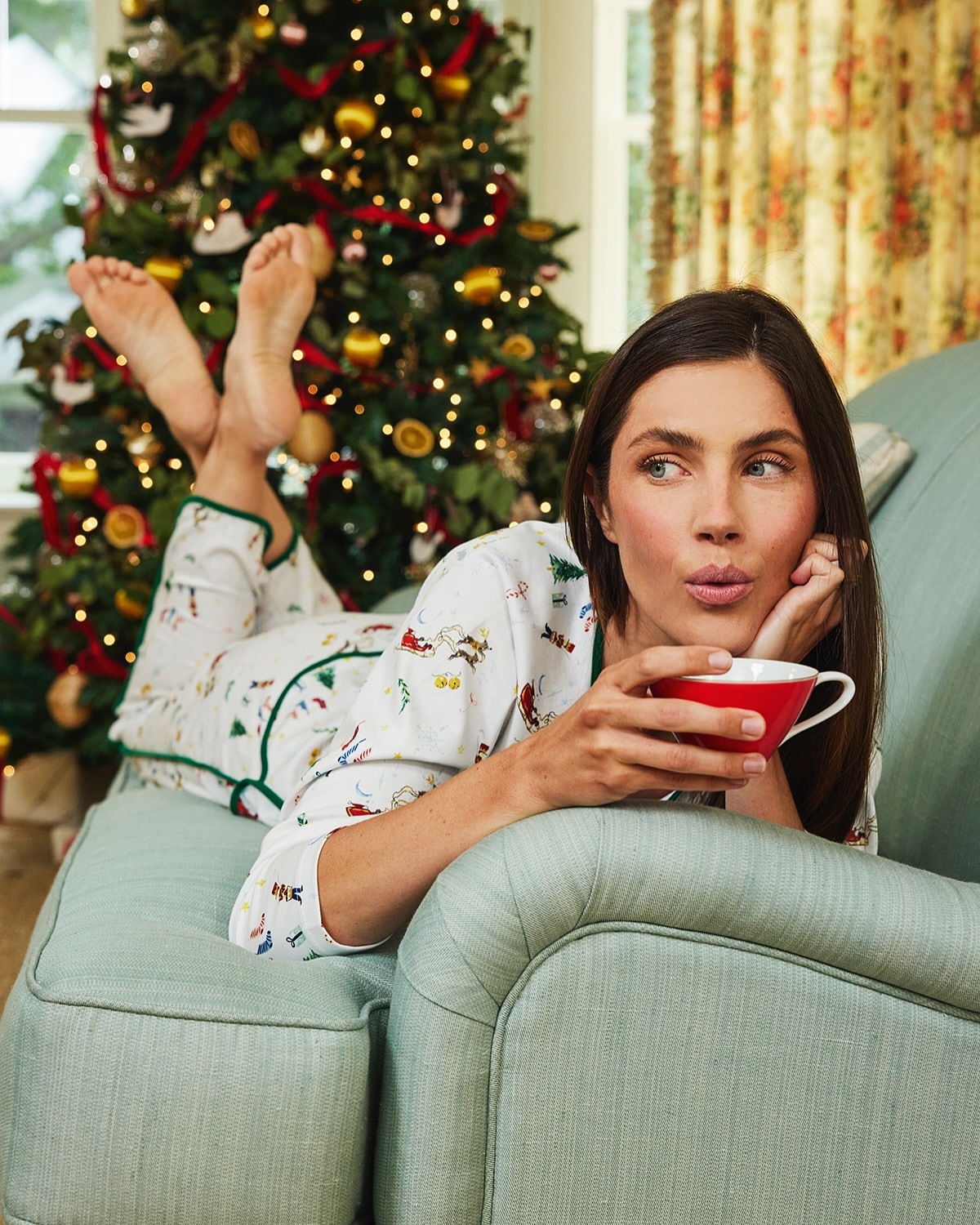 A woman in pajamas relaxing on a green chaise lounge with a mug, a Christmas tree with lights is in the background.