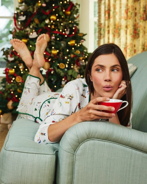 A woman in pajamas relaxing on a green chaise lounge with a mug, a Christmas tree with lights is in the background.