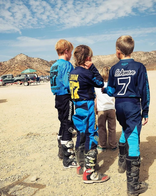 Four children wearing matching motorsport jerseys and protective boots standing on a dirt track with mountains in the distance.