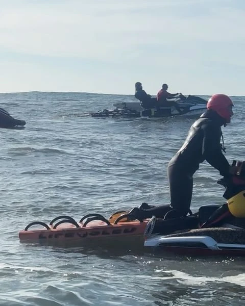 People riding personal watercraft on the ocean with rescue sleds attached. Water is choppy with a cloudy sky in the background.