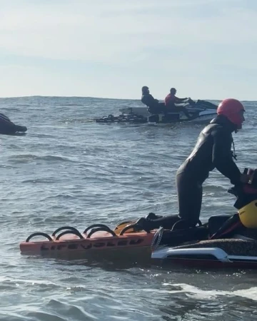 People riding personal watercraft on the ocean with rescue sleds attached. Water is choppy with a cloudy sky in the background.