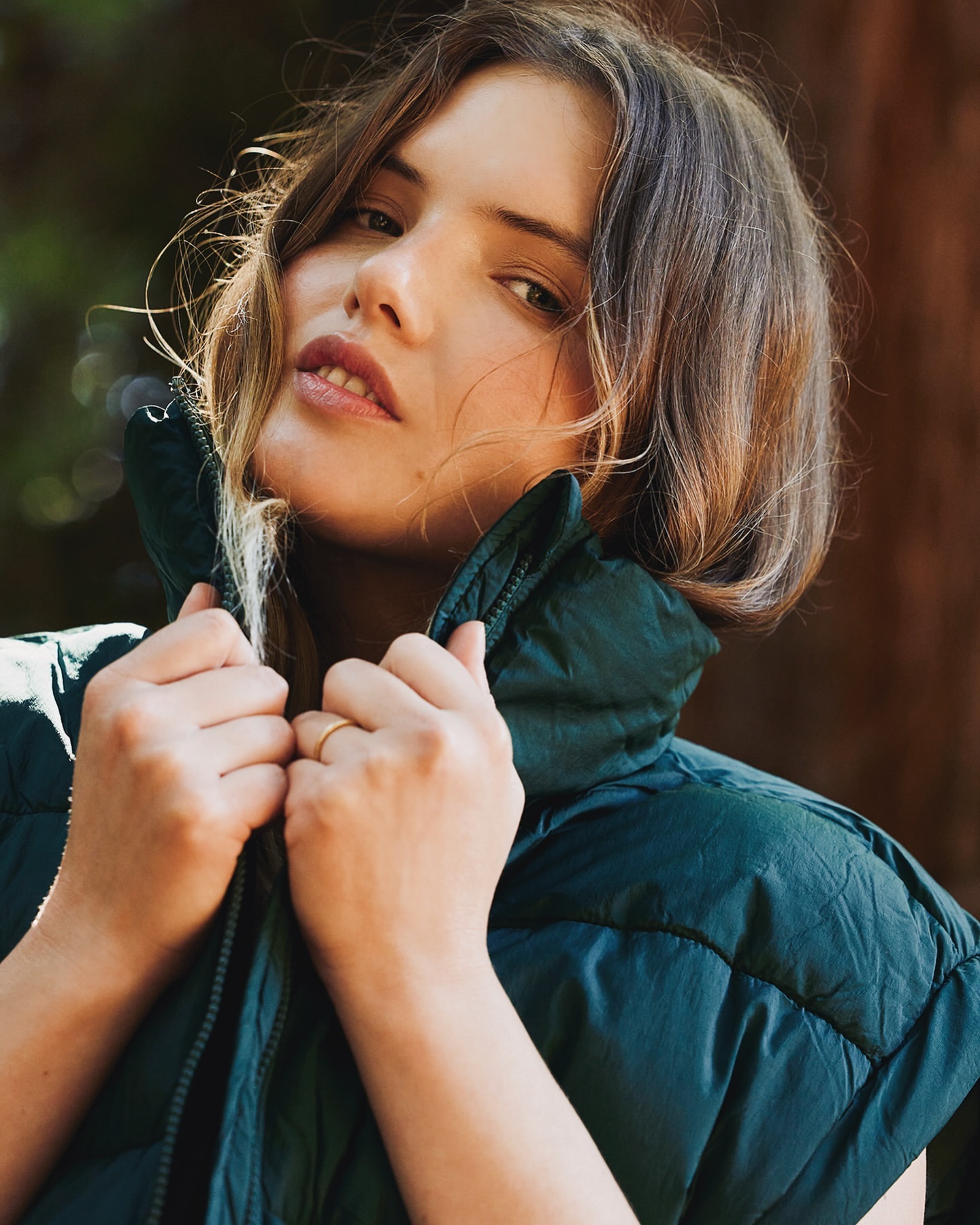 Portrait of a woman with light brown hair wearing a dark green puffer vest in an outdoor setting with blurred background foliage.