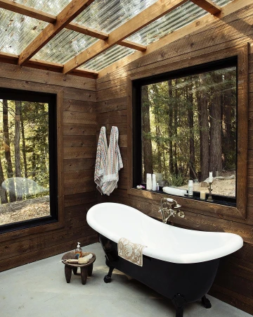 An indoor bathroom with a black and white clawfoot tub, wooden walls, and large windows looking out into a forest.