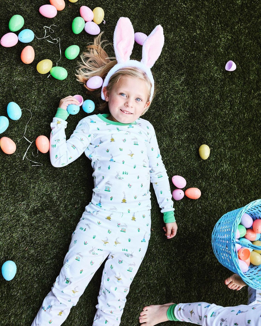 Young girl wearing bunny ears lies on artificial turf surrounded by pastel Easter eggs and a blue wicker basket filled with more eggs.