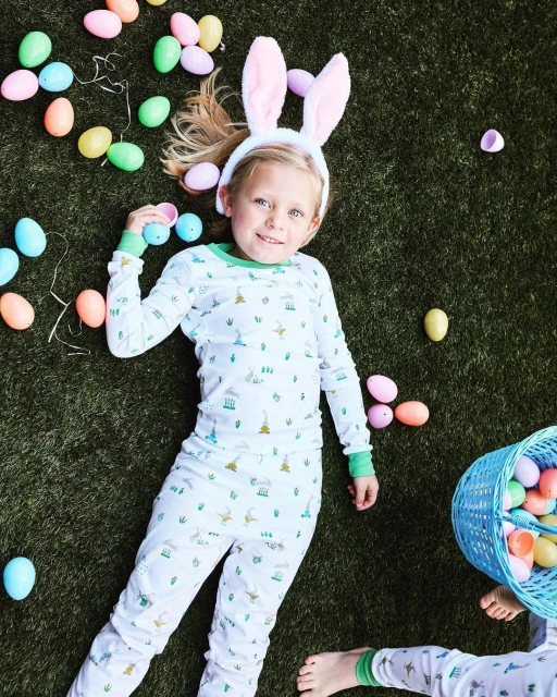 Young girl wearing bunny ears lies on artificial turf surrounded by pastel Easter eggs and a blue wicker basket filled with more eggs.