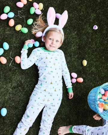 Young girl wearing bunny ears lies on artificial turf surrounded by pastel Easter eggs and a blue wicker basket filled with more eggs.