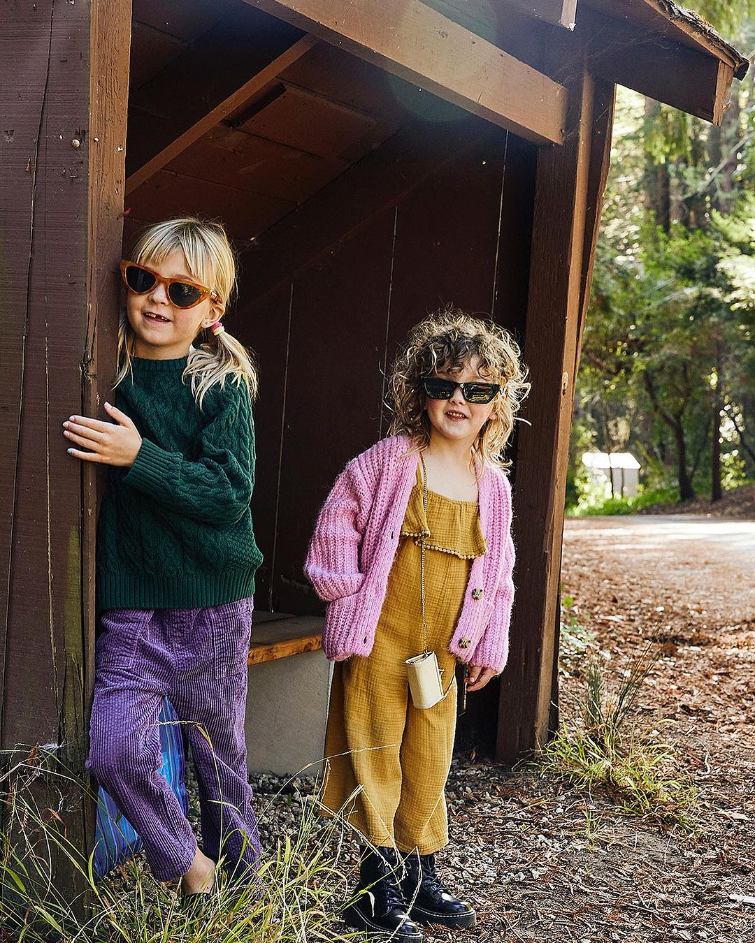 Two young girls in trendy casual outfits pose outdoors near a wood structure, with focus on fashion, eyewear, and autumn colors.