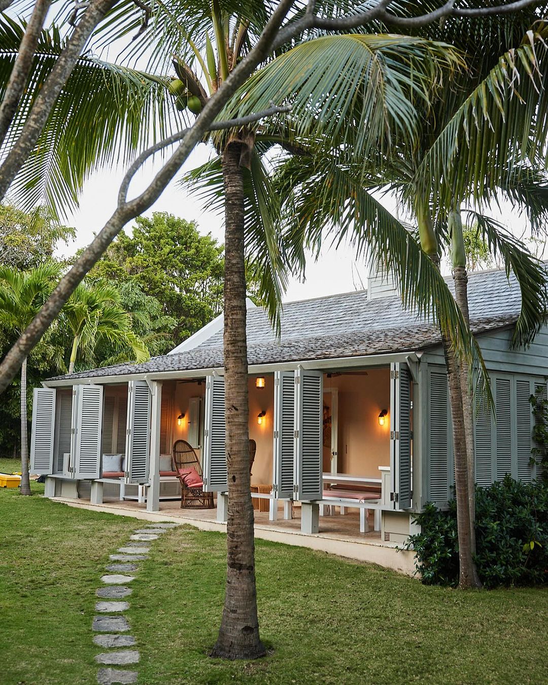 Exterior of a tropical beach house with a stone path, palm trees, and gray siding with white trim in a sunny, outdoor setting.