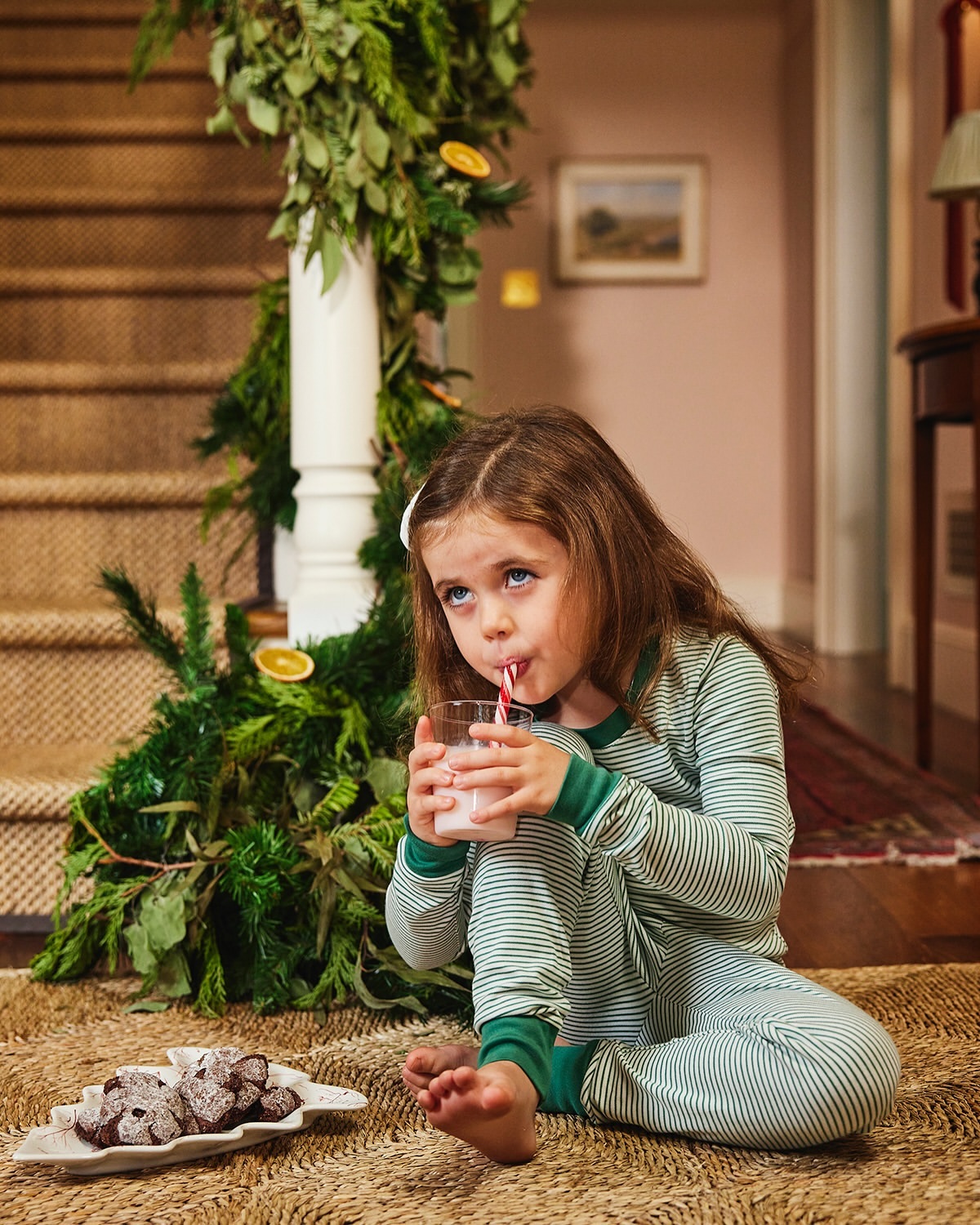 A young girl in striped pajamas sits on a rug by a staircase decorated with greenery, drinking milk and enjoying chocolate cookies.