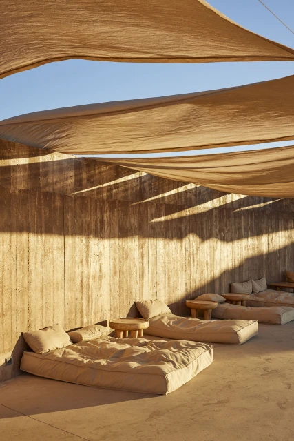 Modern outdoor lounge area with daybeds and small wooden tables under a beige fabric canopy, sunny ambiance, architectural photography.