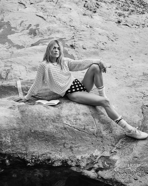 Black and white portrait of a young blonde woman sitting on a rock outdoors wearing a sweater, polka dot shorts, socks, and sneakers.