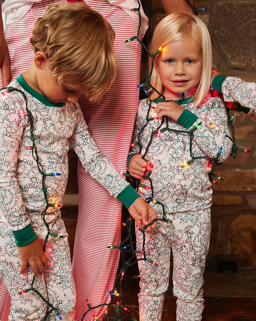 Two young children pose indoors in matching patterned pajamas wrapped in string lights, accompanied by an adult in striped pajamas.