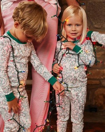 Two young children pose indoors in matching patterned pajamas wrapped in string lights, accompanied by an adult in striped pajamas.