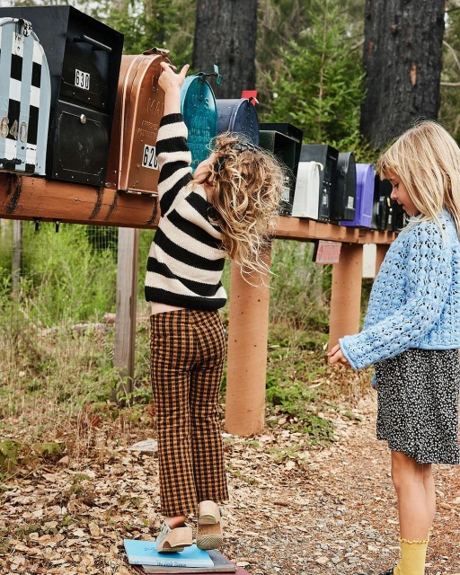 Two young girls in a wooded area looking at a row of mailboxes, one girl reaching for a mailbox; trees and greenery in the background.
