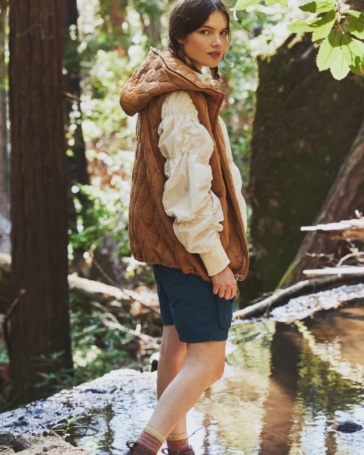 Woman wearing a quilted brown vest and dark shorts standing in a forest over a stream in a medium shot with natural light.