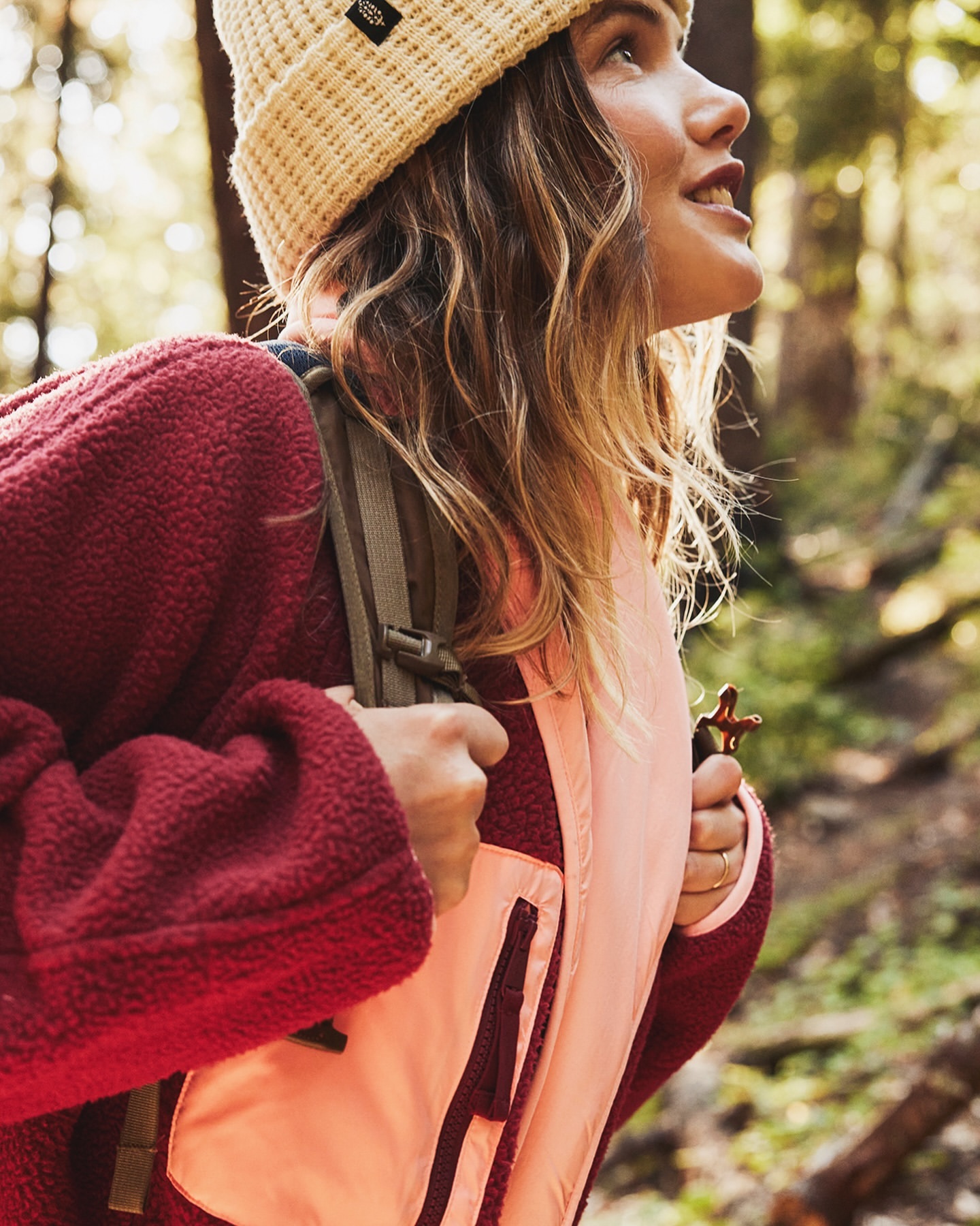 A woman hikes in a sunlit forest, wearing a beanie, fleece jacket, and backpack, looking upward with a smiling expression.