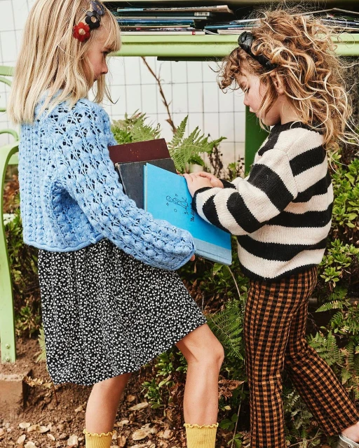 Two young girls outside holding books. One wears a blue crocheted sweater and floral dress; the other wears a striped sweater and checkered pants.