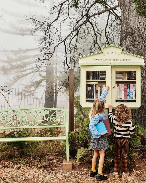 Two children selecting books from a little free library, standing outdoors on a path near a bench surrounded by greenery and trees.