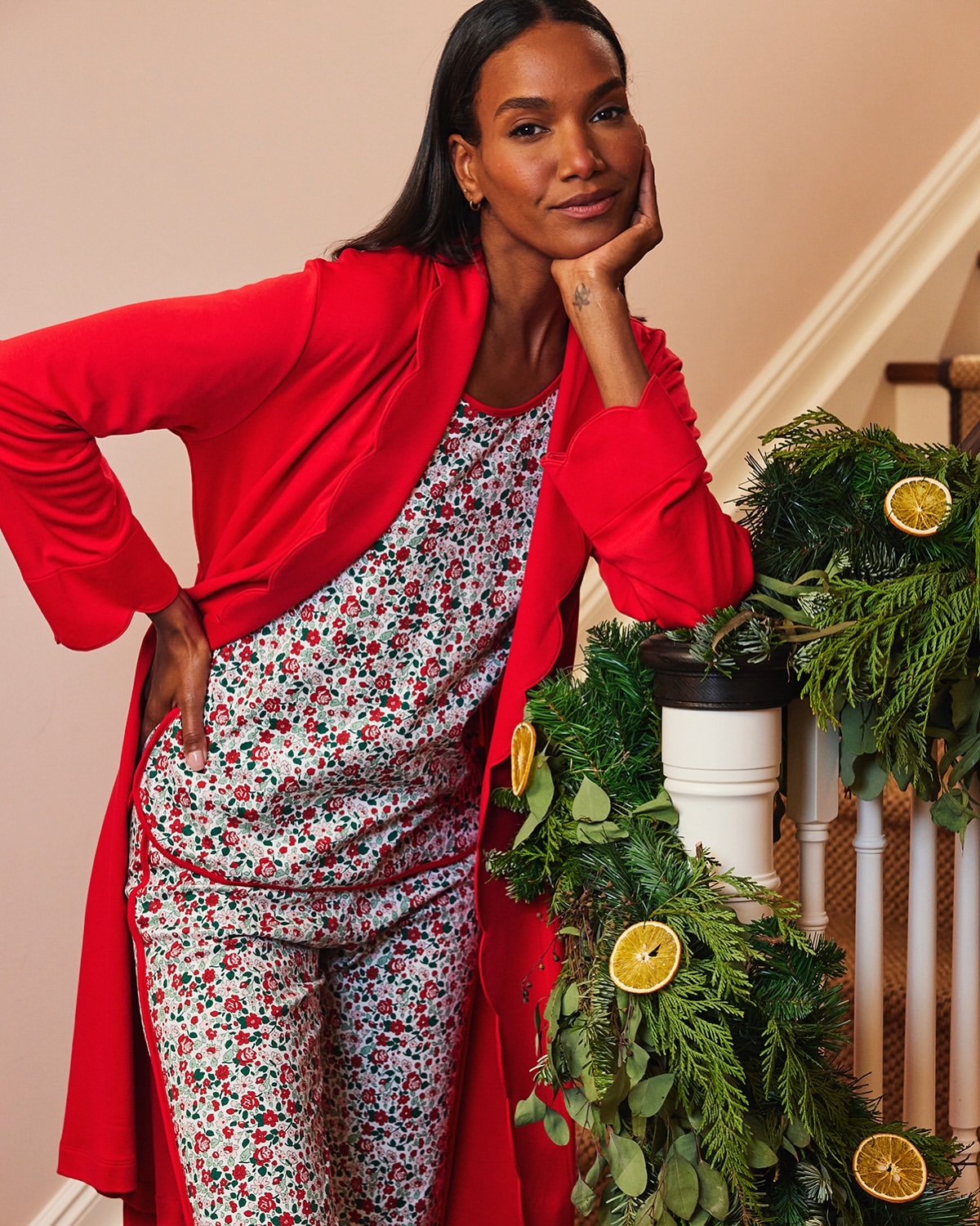Elegant dark-haired woman in red robe and floral pajamas leaning on staircase banister with festive evergreen garland and orange slices.